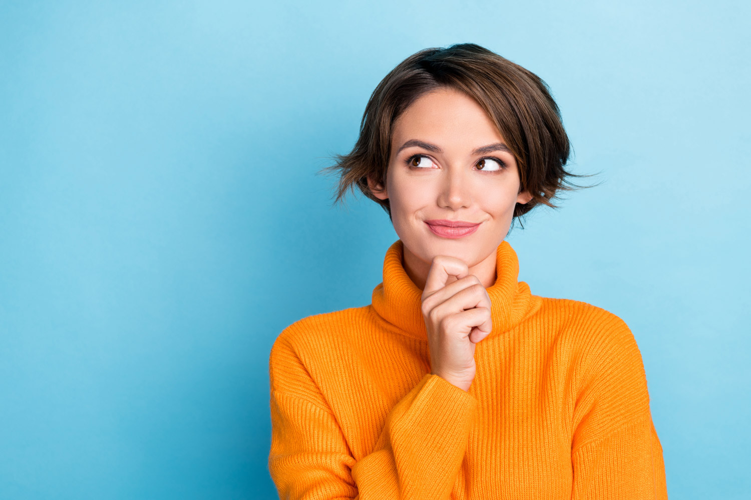 A woman in an orange turtle neck and her hand to her chin, smiles playfully